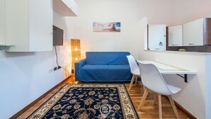Een kleine kamer met een blauwe bank, een wit bureau, een kamerkleed met dessin en een tv aan de muur.