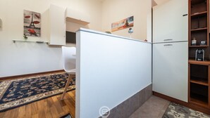 Een kleine keuken met een witte koelkast, een magnetron en een klein kastje. Er staat een stoel en een kleed in de kamer.