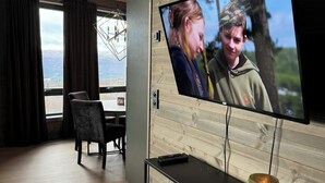Una sala moderna con un televisor grande montado en una pared de madera, una zona de comedor con mesa y sillas, y vistas al exterior a través de amplias ventanas.