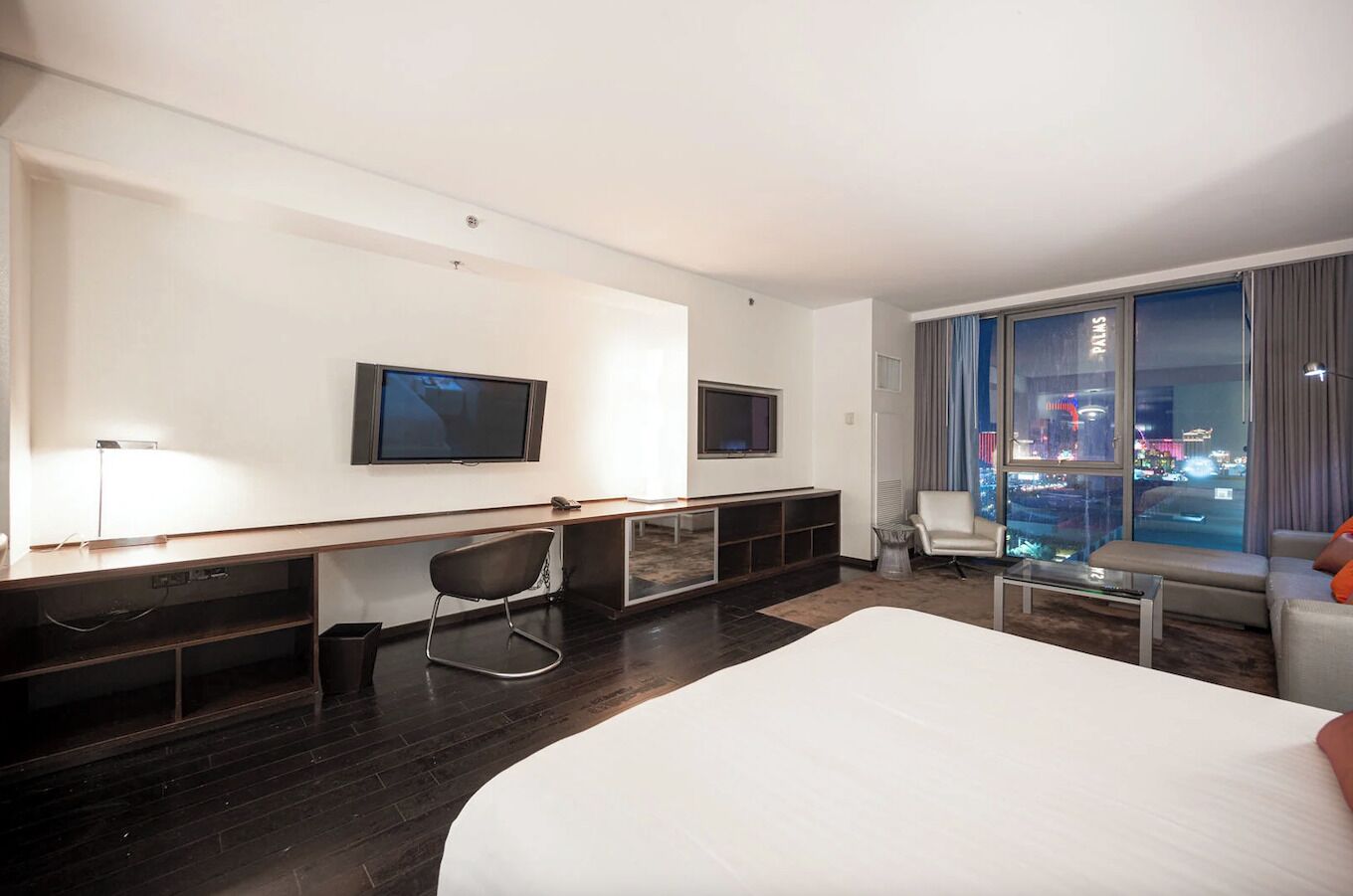 A modern hotel room with a large bed, a desk with a chair, a TV mounted on the wall, and a view of a cityscape through large windows.