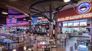 Interior de un restaurante con varias mesas y sillas, un letrero de neón que dice 'Earl of Sandwich' y una barra con productos alimenticios.