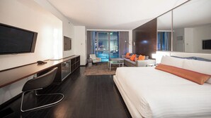 Una habitación de hotel moderna con una cama grande, un escritorio con televisor, una zona de estar con sofá y sillones, y vistas a la ciudad a través de amplios ventanales.