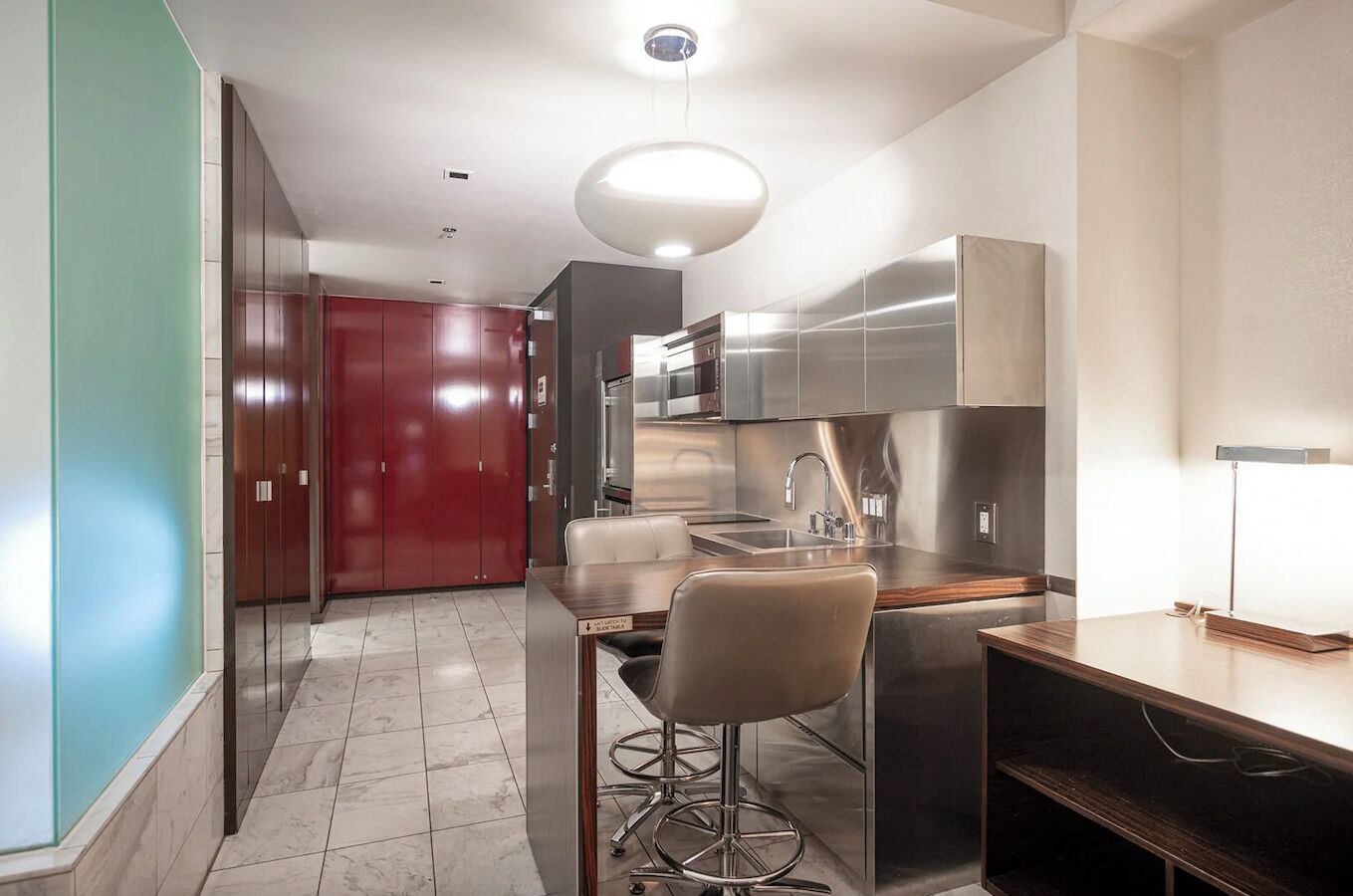 A modern kitchen with stainless steel appliances, a bar stool, and a red door in the background.