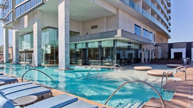A hotel pool area with lounge chairs, a clear pool, and a modern building with large windows and a stone pillar. - Palms Luxury Strip View 1BD/1.5BA Open Balcony#29KA (Las Vegas)