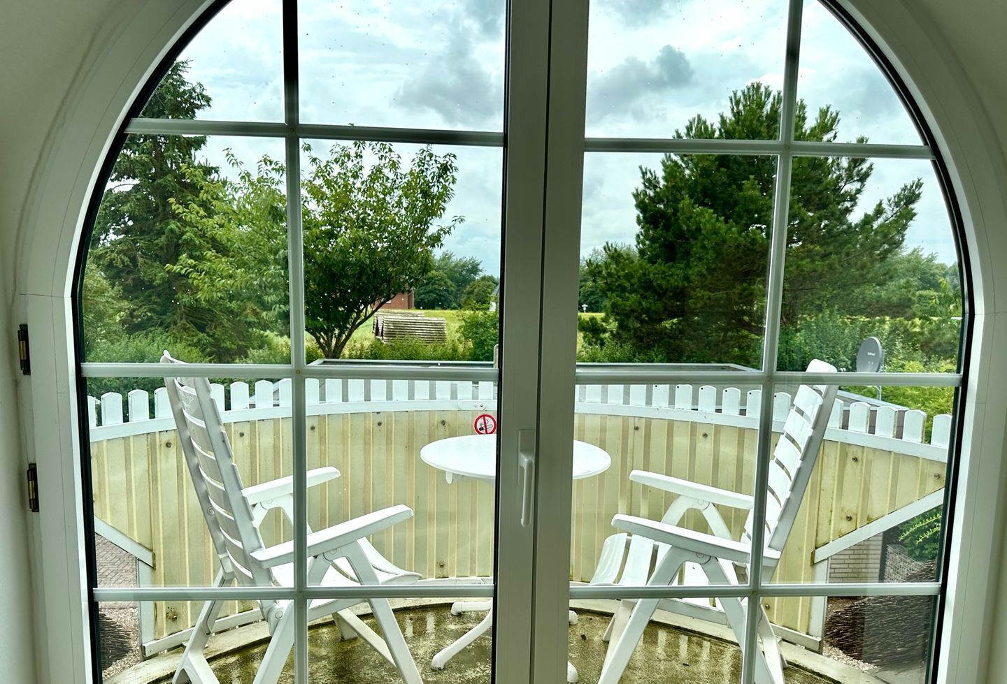 A view from a room with a large arched window overlooking a wooden deck with white chairs and a small table.