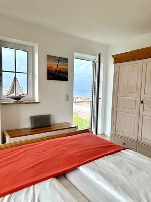 A bedroom with a bed, a desk, a chair, a wardrobe, and a view of the beach through the window.