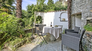 An outdoor patio with wicker furniture, a table with a white tablecloth, and a bottle of wine. - Romantic Terrace With Lake View (Lugano)