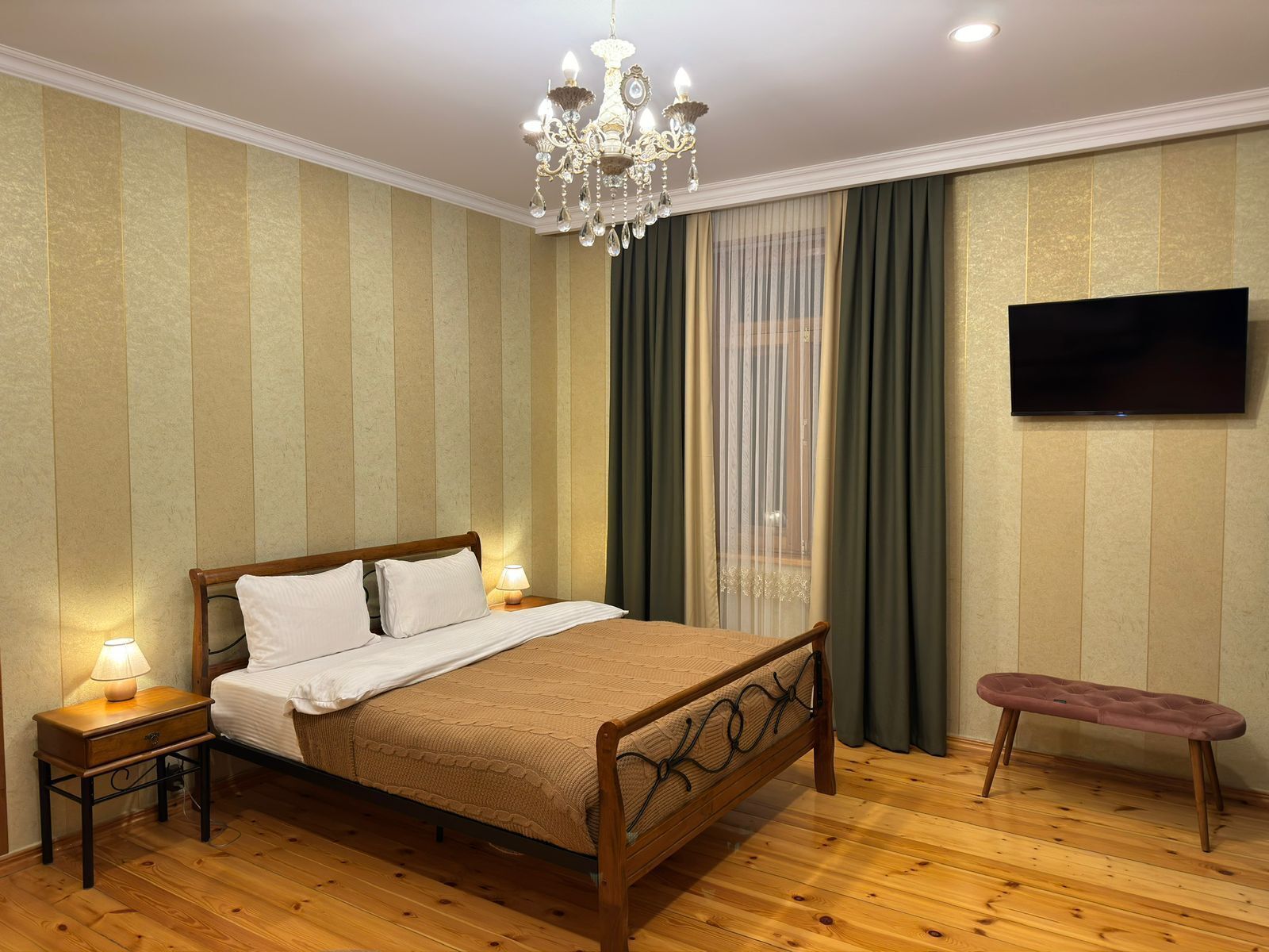 Un dormitorio con cama de madera, una mesita de noche con lámpara, un banco, una araña y televisor, además de papel tapiz a rayas.