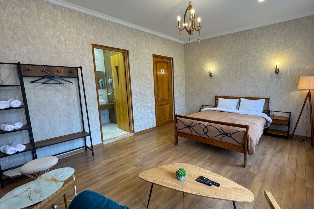 Ein Hotelzimmer mit einem hölzernen Bett, einem kleinen Tisch mit Fernbedienung, einem Sessel, einem Regal mit Handtüchern und einem Spiegel.