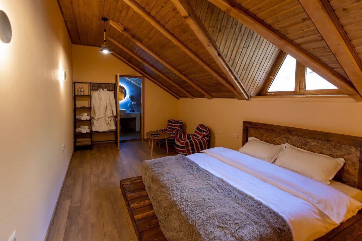 Ein Schlafzimmer mit Holzbalkendecke, einem Bett mit weißen Leinentüchern, Holzboden und einem Fenster, das natürliches Licht hereinlässt.