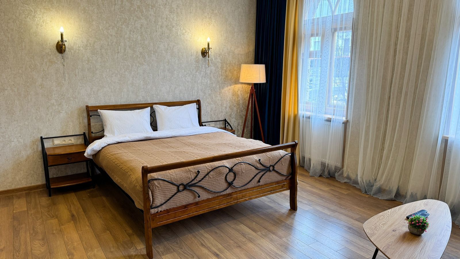 Ein Schlafzimmer mit einem hölzernen Bett, Nachttischen, einer Lampe und Blick ins Freie durch leichte Vorhänge.