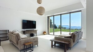 Ein modernes Wohnzimmer mit Flachbildfernseher, zwei Sofas, einem Couchtisch und einem großen Fenster mit Blick ins Freie.