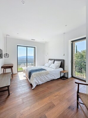 A bedroom with a large bed, wooden flooring, a small table, and a view of the outdoors through a large window.