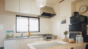 A compact kitchen with white cabinets, a gas stove, a microwave, and a refrigerator. There's a window above the sink and a clock on the wall. - CU Host CASA (Osaka)