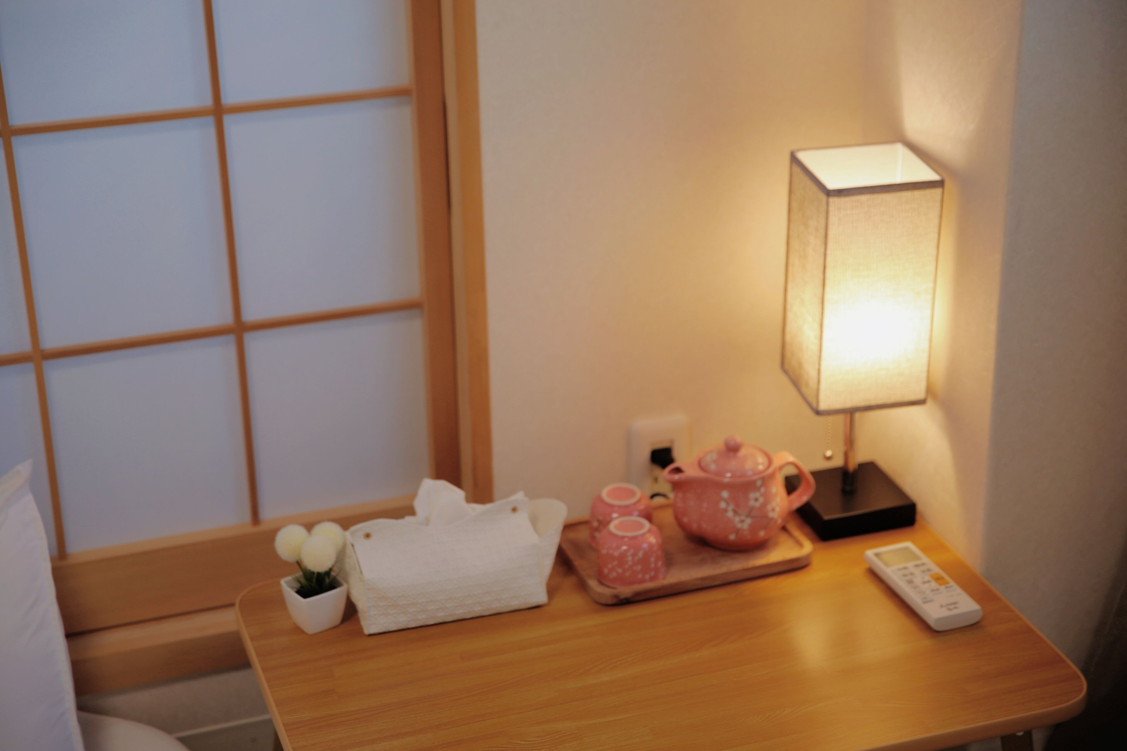 A neatly arranged bedside table with a remote control, a pink teapot, a white tissue box, and a small potted plant.