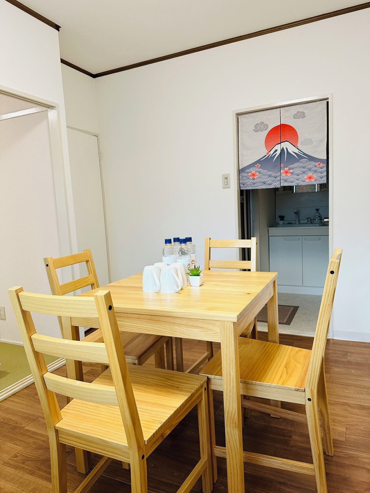 A dining area with a wooden table and chairs, a small potted plant, and a wall-mounted artwork depicting a mountain and sun.