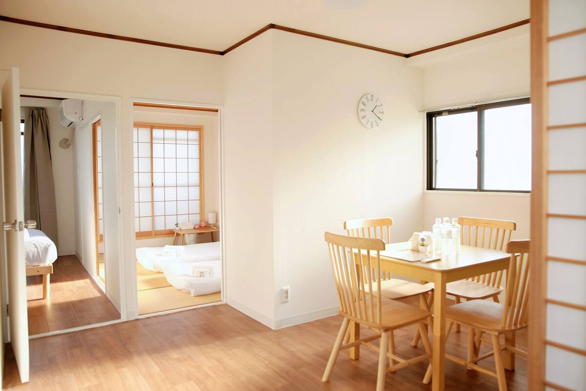 Una habitación con piso de madera, una mesa de comedor con sillas, una cama, un ventanal y un reloj en la pared.