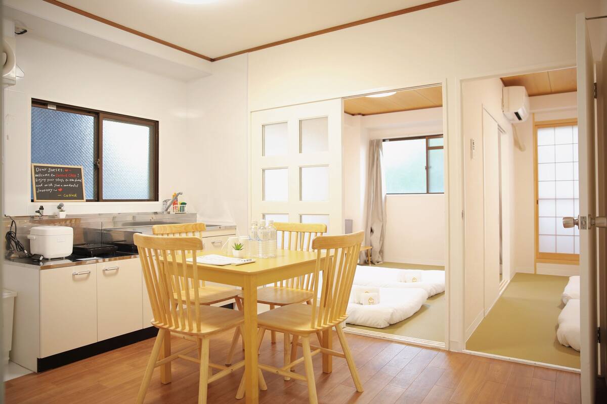 Habitación de hotel compacta con kitchenette, zona de comedor con sillas de madera, baño pequeño con perchero y puerta corredera que da a un balcón.