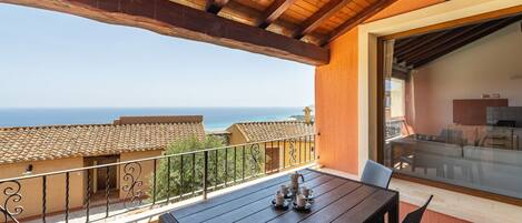 A balcony with a wooden table and chairs, a view of the sea, and a glass door leading to an indoor area.