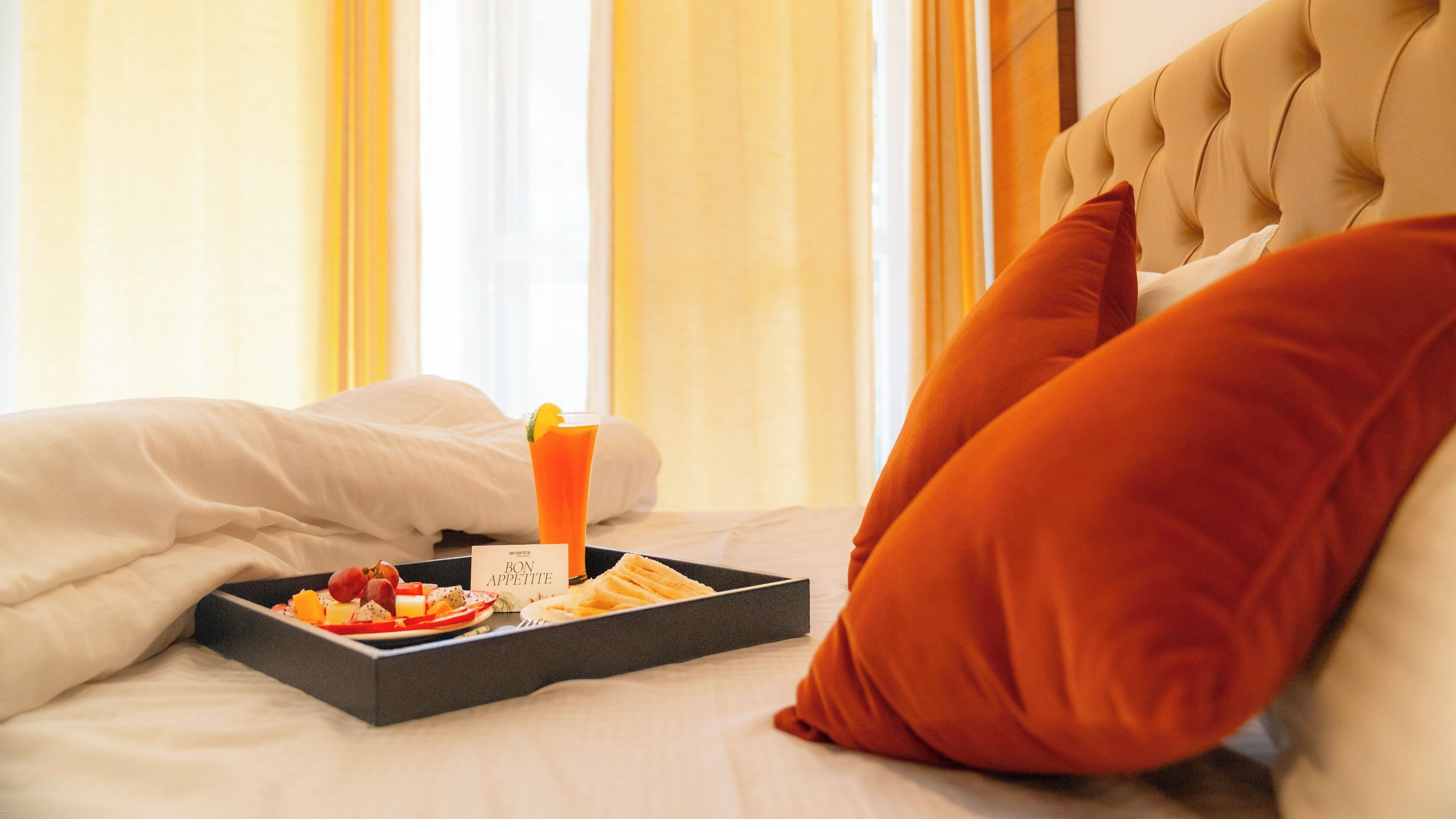 Cama bem arrumada com uma bandeja de itens de café da manhã, incluindo torradas, frutas e um copo de suco de laranja, em um quarto de hotel.