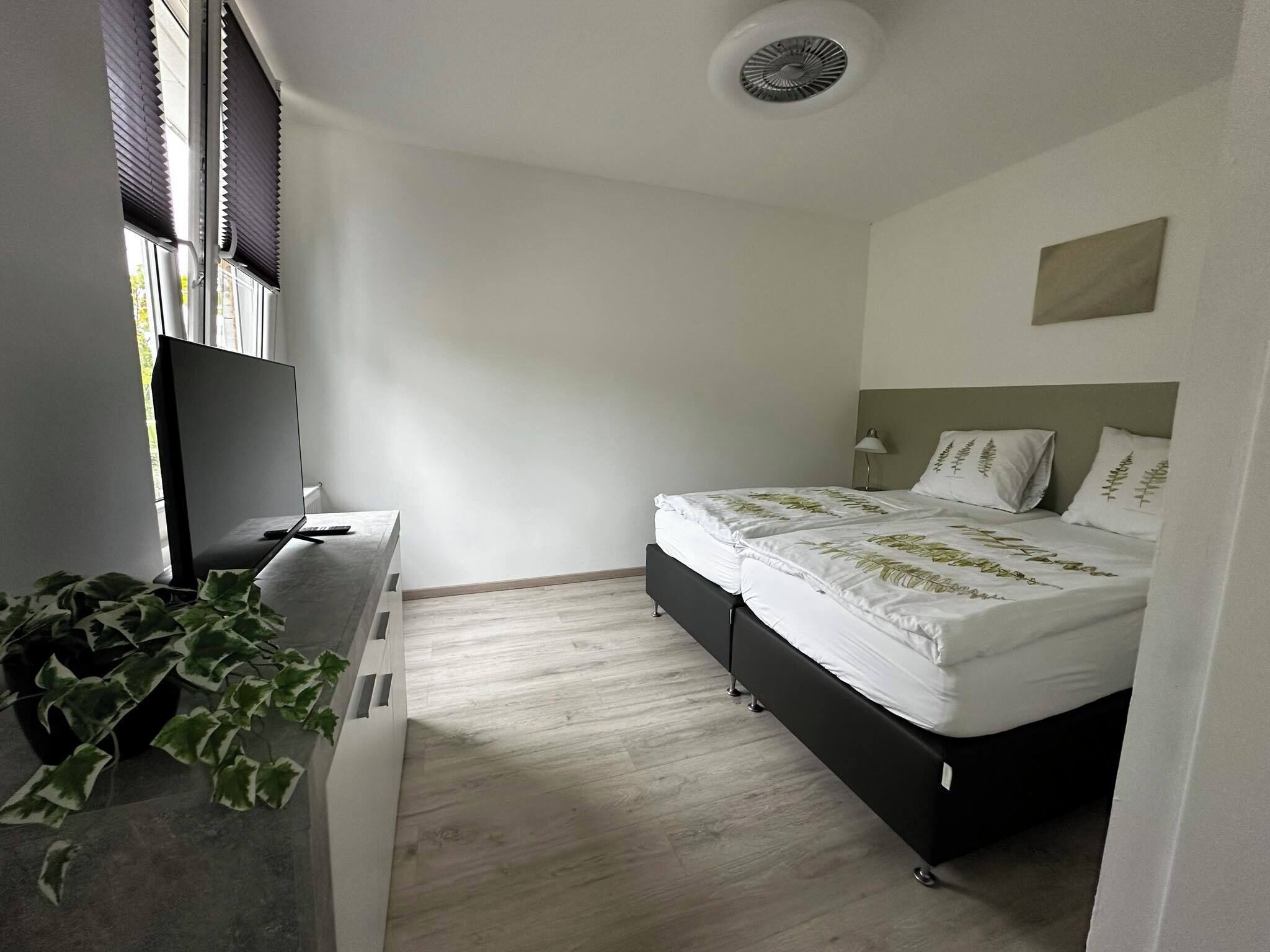 A bedroom with a bed, a desk, a TV, and a plant.