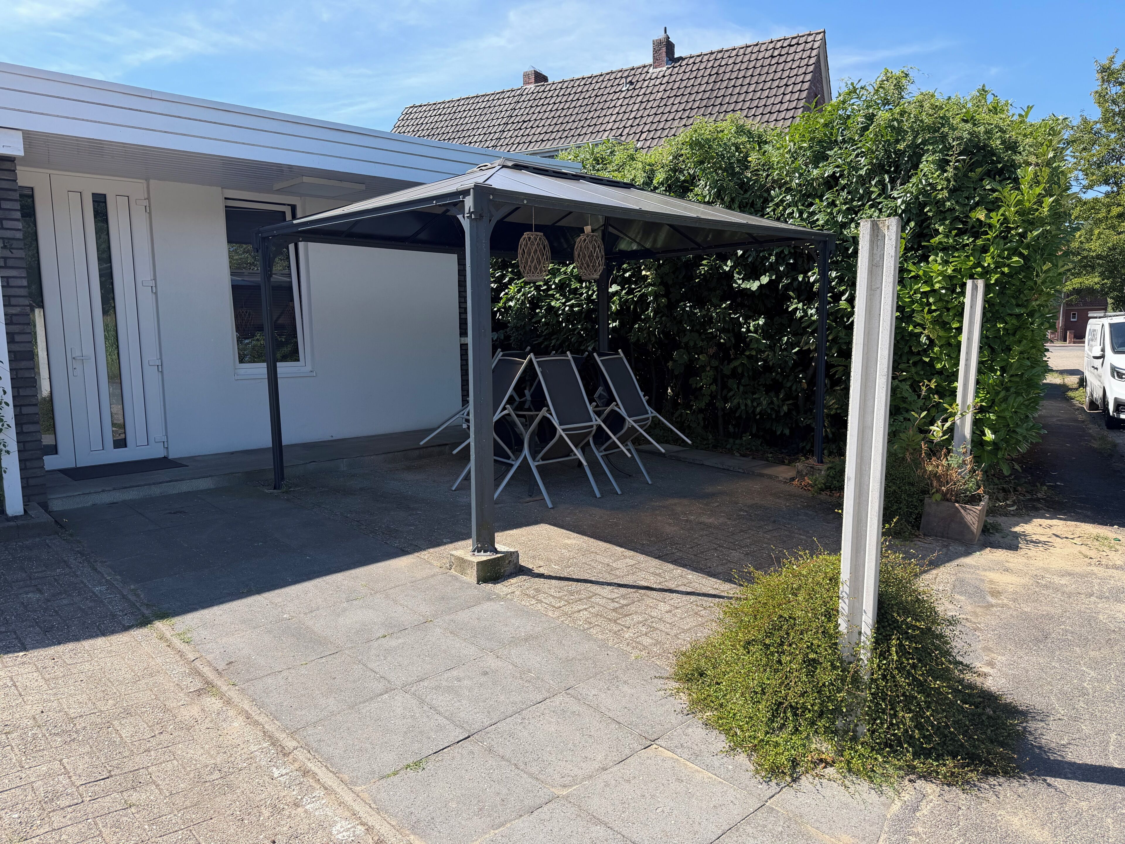 A covered outdoor area with a metal structure, a white building with a tiled roof, and a paved driveway.
