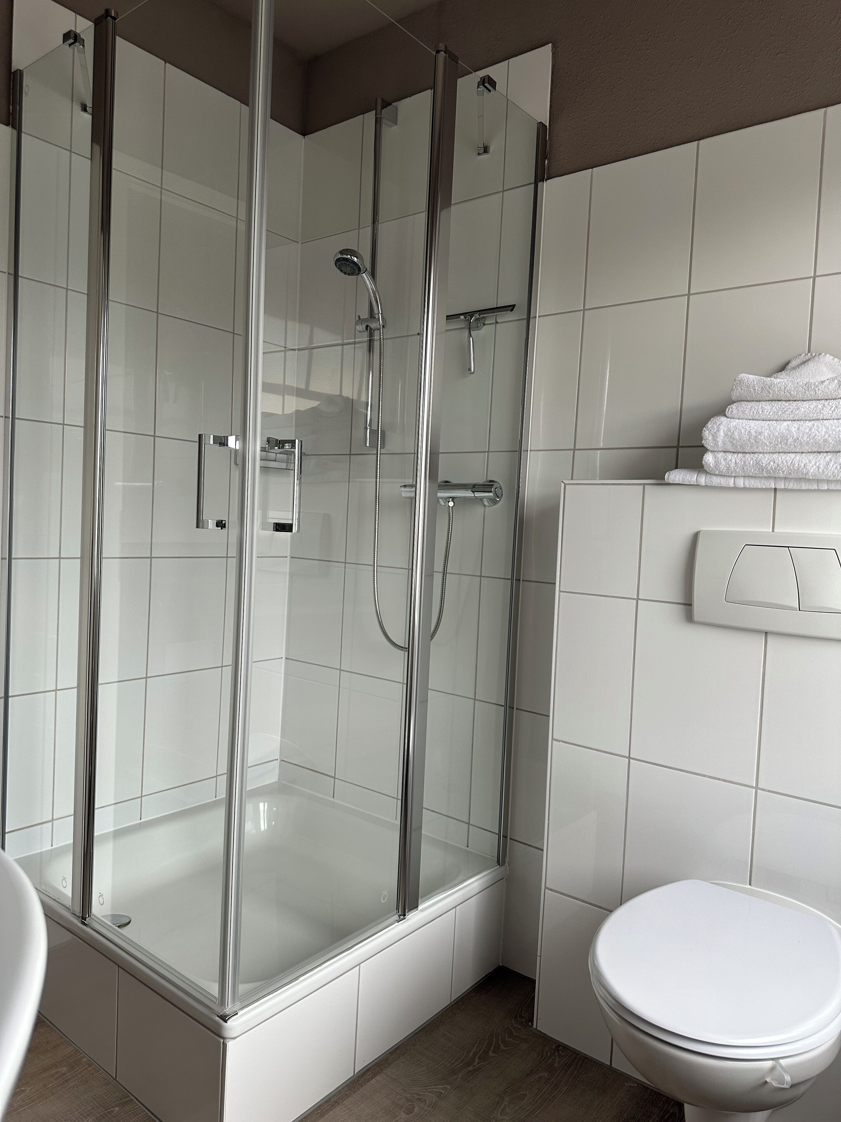 Et moderne badeværelse med bruseniche i glas, toilet og en stak hvide håndklæder.