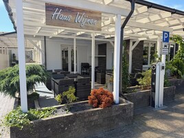 A covered outdoor seating area with wicker furniture, a sign reading 'Haus Wijn,' and a parking sign indicating a charging station.