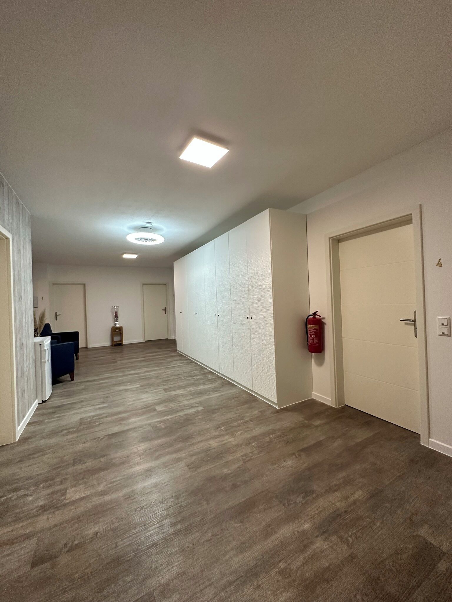 A hallway with wooden flooring, white walls, and a fire extinguisher mounted on the wall.