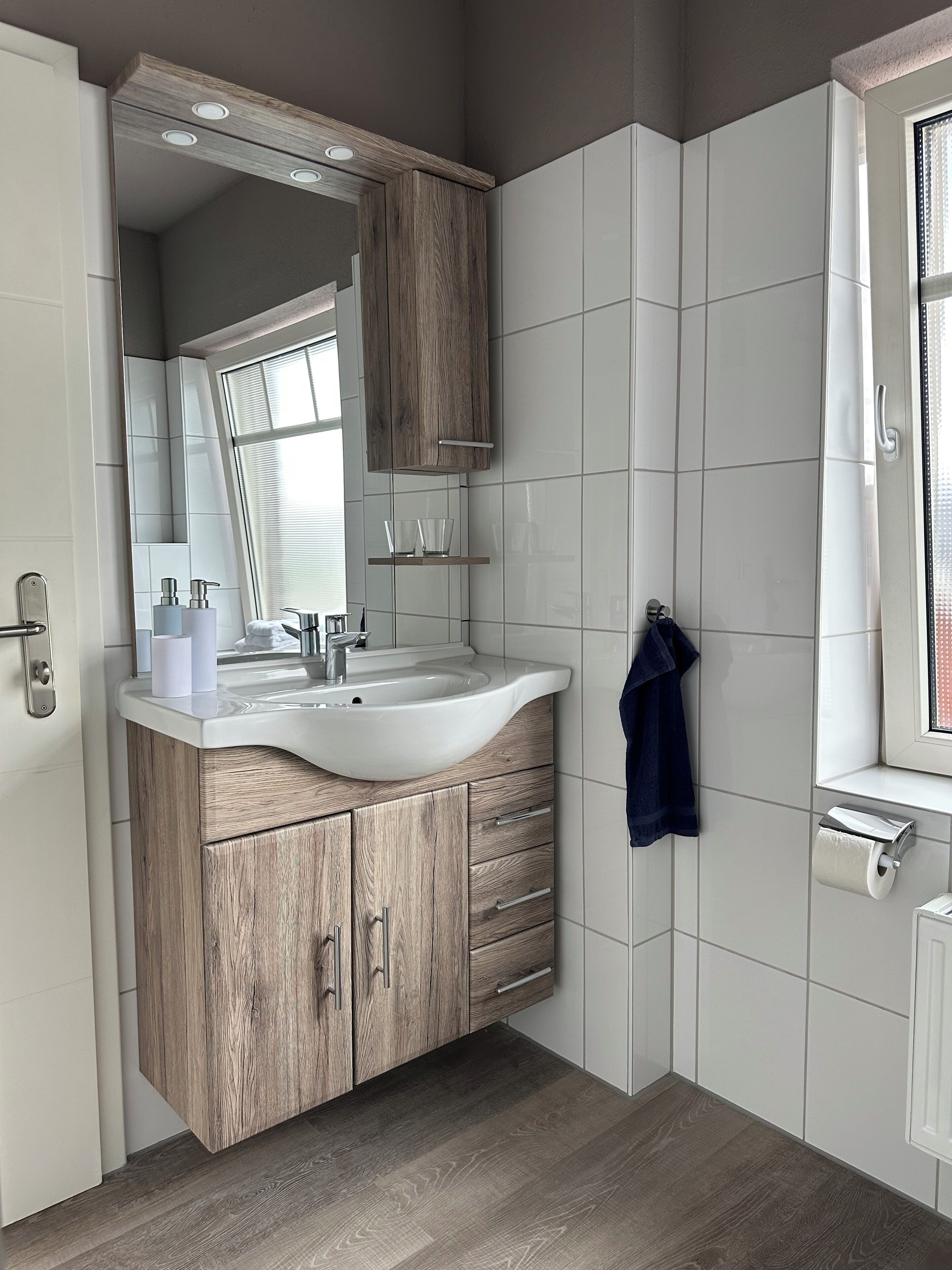 A modern bathroom with a wooden vanity, white tiles, a large mirror, and a towel hanging on the wall.