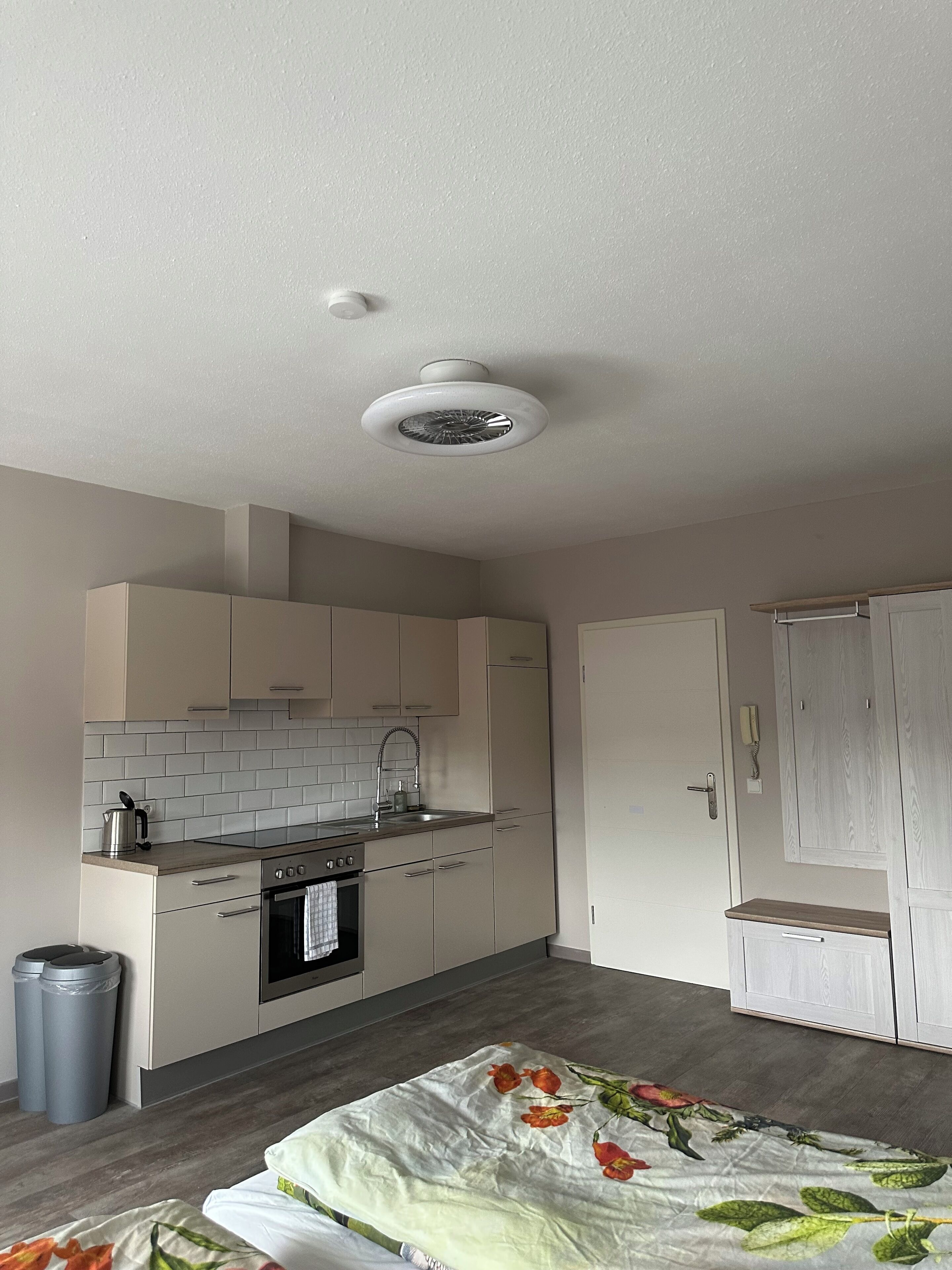 A compact kitchen with a stove, sink, and cabinets. There is a trash bin and a bed with a floral bedspread.