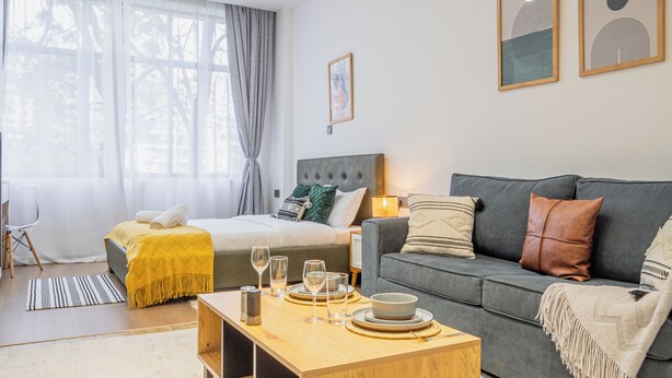 Habitaci ó d'hotel moderna amb un llit, un sofà gris, una taula de centre de fusta i una finestra gran amb cortines.