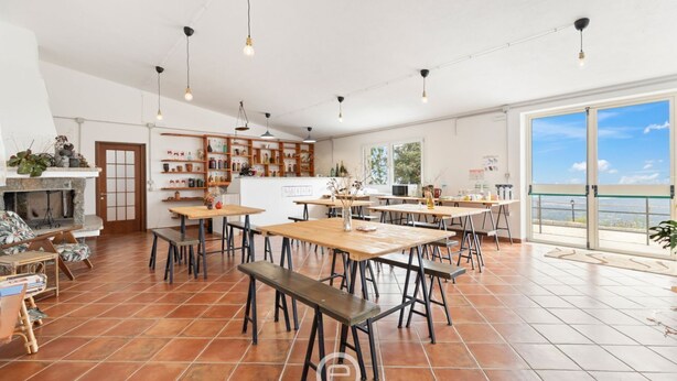Un comedor amplio con varias mesas y bancos, zona de cocina con armarios de madera y grandes ventanales que ofrecen vistas.