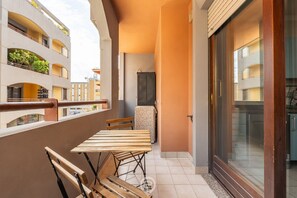 Un balcon avec une table et des chaises en bois, un réfrigérateur et une porte-fenêtre coulissante donnant sur un autre balcon avec vue sur d’autres immeubles.