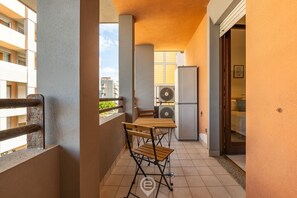 Un balcon avec une table et des chaises, un réfrigérateur et une fenêtre donnant sur un immeuble.