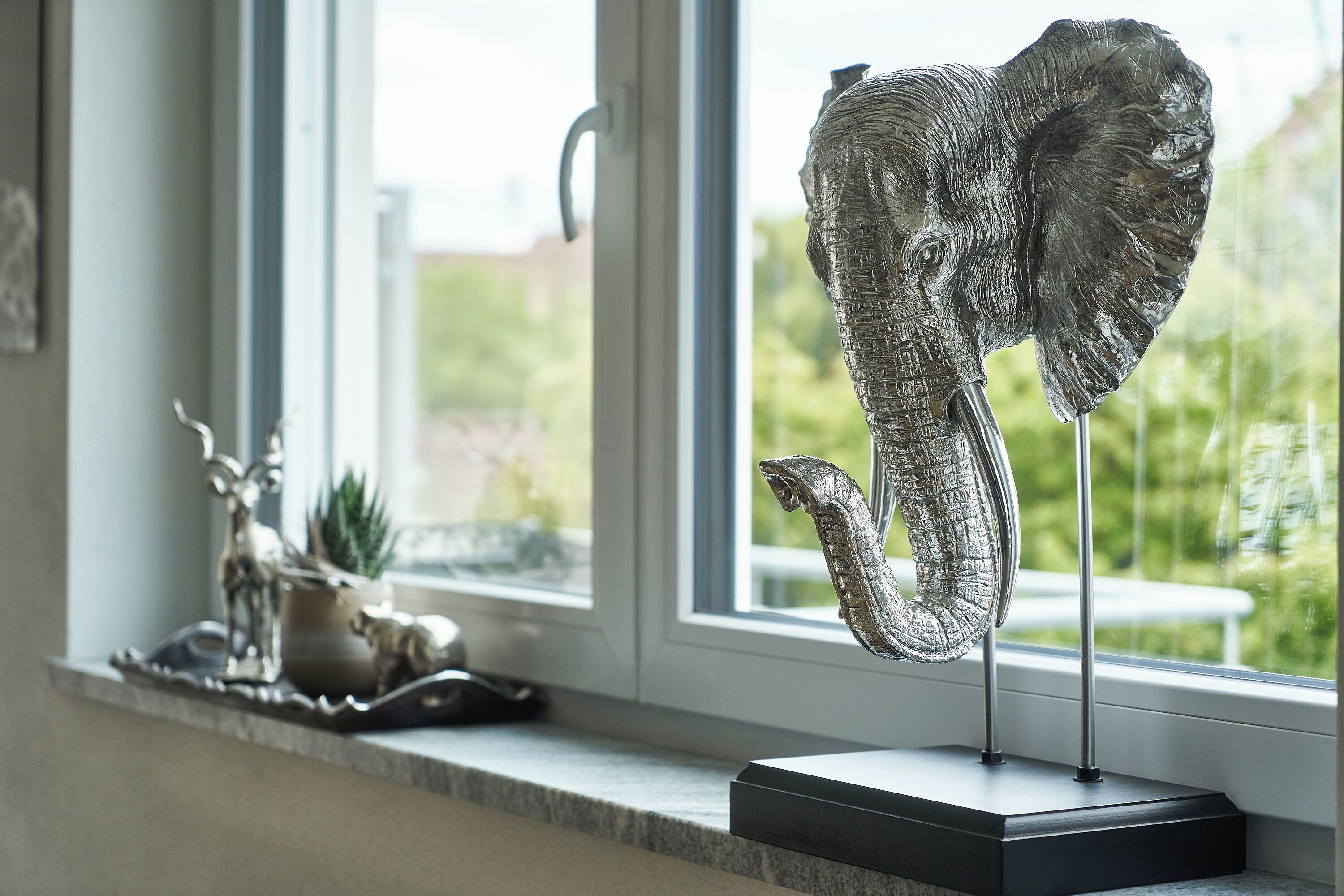 A sculpture of an elephant's head on a stand by a window with a view of greenery.