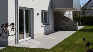Ein modernes, zweistöckiges Gebäude mit großen Fenstern, einem überdachten Balkon und einer Steintreppe, die zum Eingang führt.