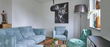 A living room with a blue sofa, a green armchair, a round coffee table with a plant, a painting of a cat, and a floor lamp.