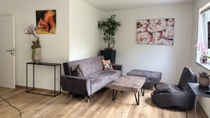 A modern living room with a grey sofa, a wooden coffee table, a grey bean bag, and a black TV stand. There is a painting of a fox on the wall and a potted plant on the coffee table. - Vacation apartment Squirrel (Günzburg)