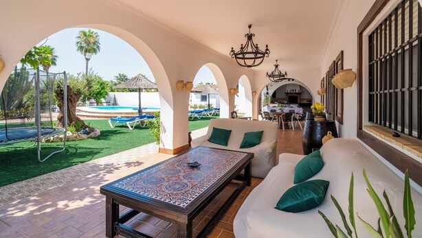 A sunlit patio with a white sofa, a patterned table, and a view of a pool and garden. - Holiday Home La Perla (La Puebla de Cazalla)