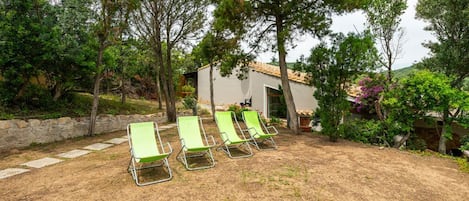 A backyard with four green lounge chairs, a stone wall, and a small building with a tiled roof.