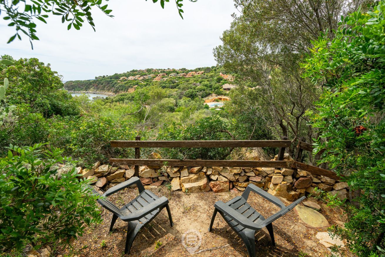 A patio with two chairs and a wooden table, surrounded by a stone wall and lush greenery, overlooking a hilly landscape with buildings in the distance.