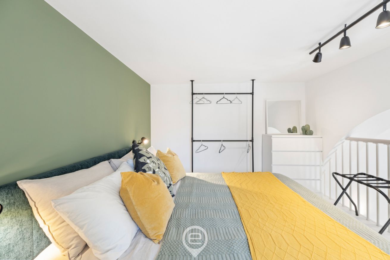 A bedroom with a bed, pillows, a bedside table, a hanging clothes rack, and a staircase.
