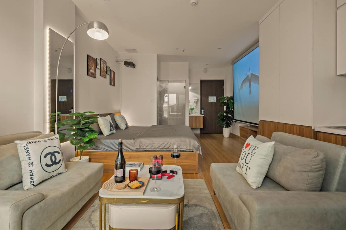 A modern hotel room with a bed, two sofas, a coffee table, a TV, and a large window.