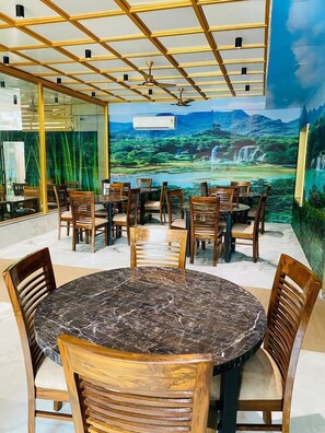 A restaurant with wooden tables and chairs, a ceiling fan, and a mural of a waterfall. - Hotel T2 Grand (Mumbai)