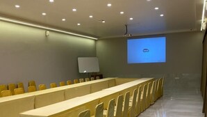 A conference room with a long table, yellow chairs, a whiteboard, and a projector screen displaying a logo. - Hotel T2 Grand (Mumbai)