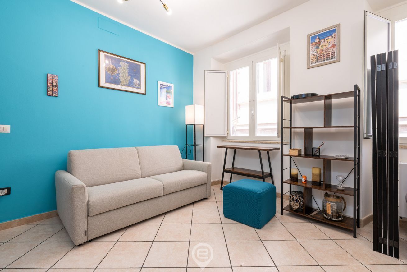 Un salon avec un mur bleu, un canapé beige, un petit pouf bleu, une table d’appoint en bois et une étagère noire contenant divers objets.