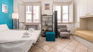 Una camera d'albergo con un letto, una sedia, un tavolino e uno scaffale con degli oggetti.