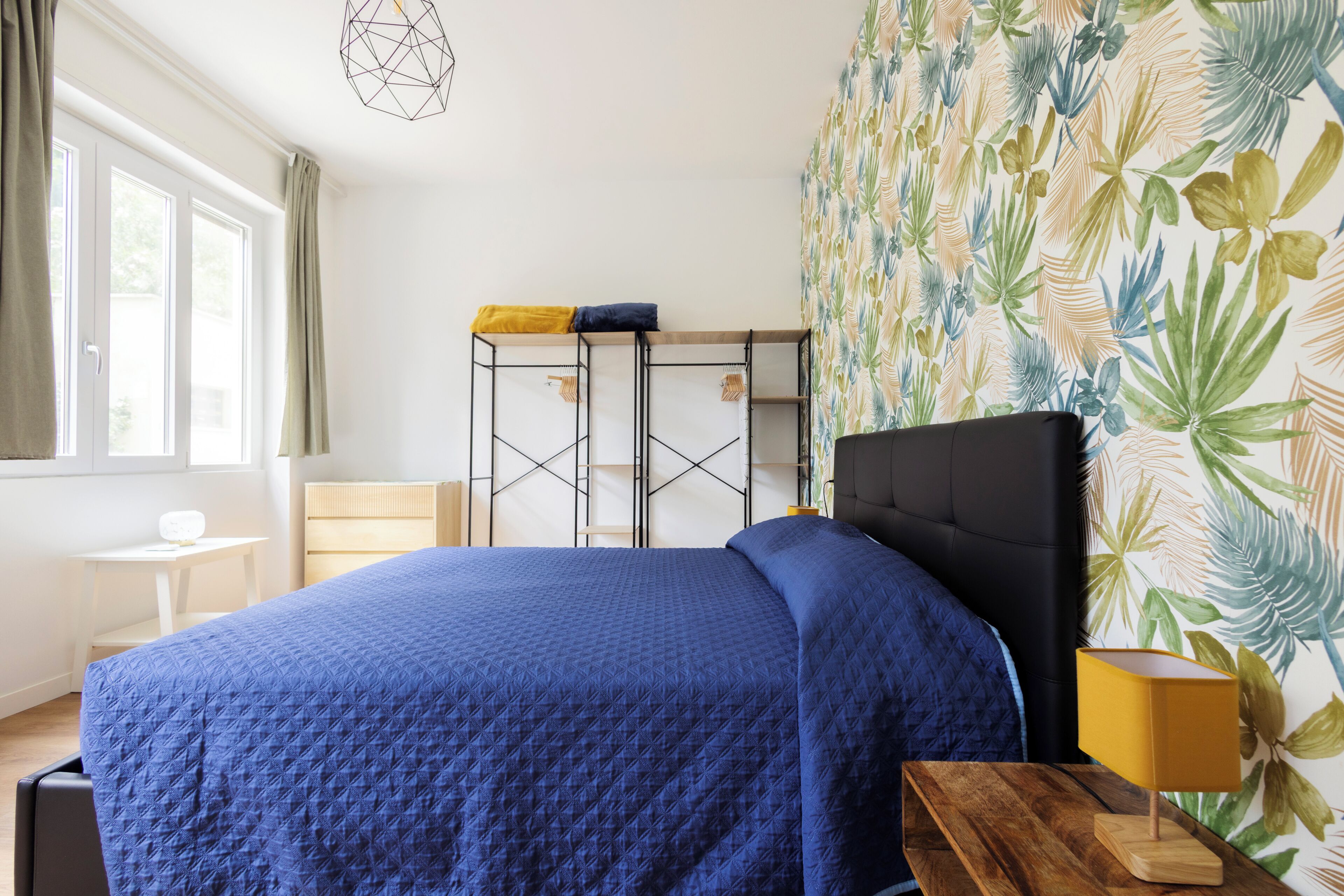 Een slaapkamer met een bed, een bureau, een raam en een muur met een tropisch bladmotief.