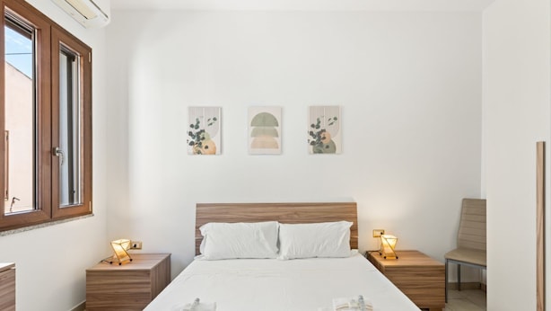 Quarto com cabeceira de madeira, uma cama grande com roupa de cama branca, duas mesas de cabeceira com abajures e três obras de arte fixadas na parede.
